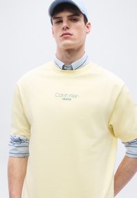 Νεαρός άνδρας που φοράει ένα ανοιχτό κίτρινο πουκάμισο Calvin Klein Jeans πάνω από ένα ριγέ πουκάμισο με κουμπιά που είναι διπλωμένο στα μανίκια και ένα ανοιχτό μπλε καπέλο του μπέιζμπολ.