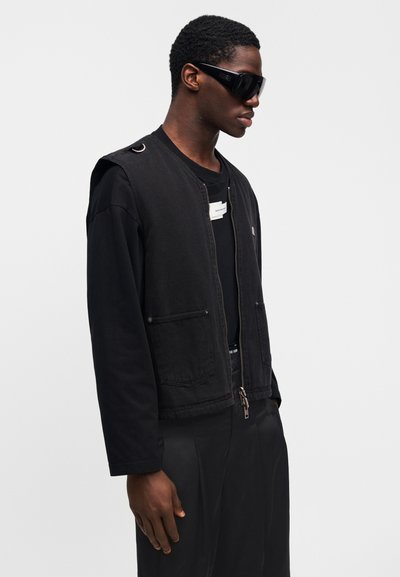 Μαύρο τζιν γιλέκο με δύο μπροστινές τσέπες και κλείσιμο με φερμουάρ, φορεμένο πάνω από μαύρο πουκάμισο με μακριά μανίκια. Διαθέτει ετικέτα στο λαιμό και oversized εφαρμογή.