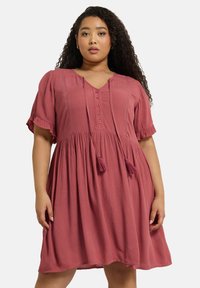 Vaaleanpunainen lyhythihainen mekko, jossa on V-pääntie, nappikiinnitys ja tupsun nyörikuvio. Kangas on tekstuurinen, kevyt ja löysä.