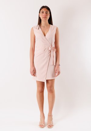 Donna in un vestito avvolgente senza maniche di colore rosa pallido con fiocco laterale, in piedi dritta contro uno sfondo chiaro e uniforme, indossando tacchi beige.