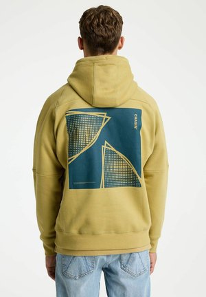 Homme portant un sweat à capuche vert clair avec des formes géométriques bleues et jaunes au dos, associé à un jean bleu clair, debout devant un fond blanc.