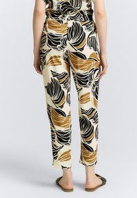 Leichte, gemusterte Hosen in Creme mit schwarzen und goldenen Blattthemen, entspannter Schnitt und verkürzte Länge, kombiniert mit Slipper-Sandalen.