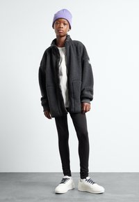Giacca in fleece nera con zip frontale, polsini a coste, abbinata a leggings neri e sneakers bianche. Un berretto viola completa l'outfit.