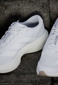 Λευκά παπούτσια τρεξίματος Adidas Adizero με παχιά σόλα τοποθετημένα σε πέτρινη επιφάνεια, που δείχνουν πλάγια και από πάνω όψη.