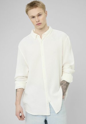 Jeune personne aux cheveux blonds courts portant une chemise blanche ample à boutons et un jean bleu clair, montrant ses avant-bras tatoués.