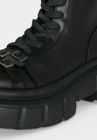 Botte de combat en cuir noir avec semelle épaisse texturée, lacets noirs et deux sangles à boucle argentées sur la zone des orteils.