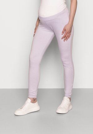 Schwangere Person trägt hellviolette Umstandsleggings und weiße Turnschuhe, steht vor einem einfarbigen Hintergrund.