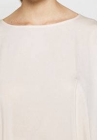Top beige chiaro realizzato in tessuto morbido e liscio, con un ampio scollo rotondo e una vestibilità che cade delicatamente. Design minimalista senza motivi visibili.