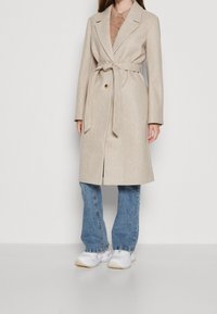 Béžový kabát z vlněné směsi s opaskem, s límci a jedním knoflíkem. Nosený přes hnědou košili a kombinovaný s modrými džíny a bílými teniskami.