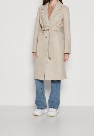 Beige Wollmischung Mantel mit einem Bindegürtel, taillierten Revers und Ein-Knopf-Verschluss. Getragen über einem braunen Hemd und kombiniert mit blauen Jeans und weißen Sneakers.