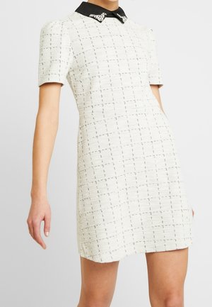 Kvinde iført kortærmet hvid tekstureret kjole med et subtilt ternet mønster og sort krave prydet med juvelbesætninger.