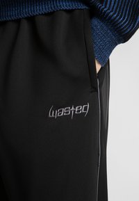 Roka v žepu, oblečen v črne hlače z vezenim belim napisom "wasted" in vidnim rokavom modrega pletenega puloverja.