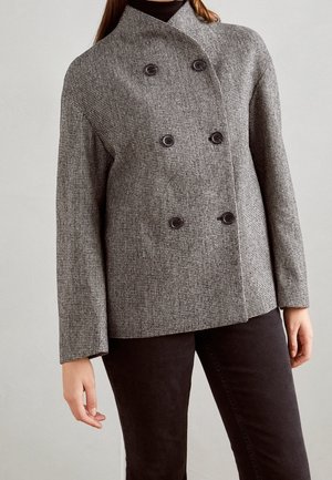 Vrouw die een grijs geruit dubbelgeknoopt jasje draagt met zes zwarte knopen, gecombineerd met een zwarte broek, tegen een effen achtergrond.