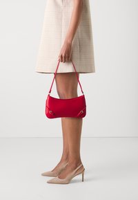 Žena v béžových lodičkách a sukni držící malou červenou kabelku za popruh na bílém pozadí.