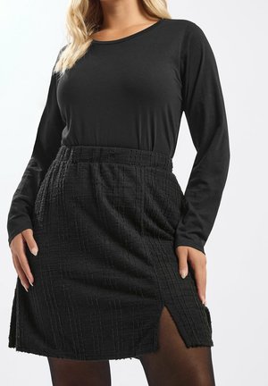 Blond vrouw met een zwart top met lange mouwen en een getextureerde zwarte rok met een kleine voorste split, staand tegen een effen achtergrond.