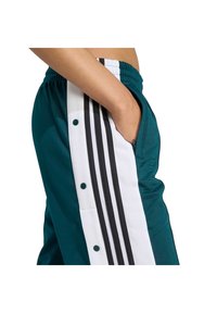 Teal športové nohavice s bielymi bočnými panelmi a čiernymi pruhmi, so zapínaním na gombíky a vreckami, vyrobené z hladkého, ľahkého materiálu.