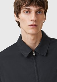 Zwarte, met rits sluitende jas met een spitse kraag, met een gladde textuur en minimalistisch ontwerp, gedragen door een persoon met kort, tousled haar.