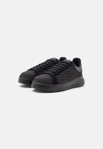 Paire de baskets basses noires avec lacets noirs, semelles épaisses rainurées et texte à motif sur la tige en tissu.