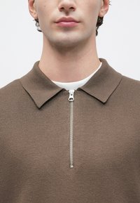 Primer plano de una persona que lleva un suéter de punto marrón con cremallera y cuello sobre una camisa blanca, con la cremallera parcialmente cerrada en el cuello.