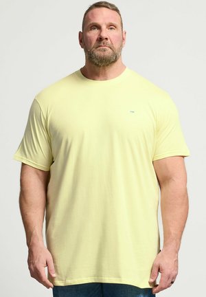 Uomo muscoloso con la barba, in piedi di fronte, indossa una maglietta giallo chiaro a maniche corte semplice e pantaloni scuri su uno sfondo neutro.