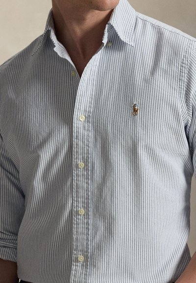 Άνδρας που φοράει μπλε και λευκό κάθετα ριγέ πουκάμισο με μικρό κεντημένο λογότυπο στο αριστερό μέρος του στήθους, με το γιακά ξεκουμπωμένο.