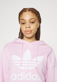 Rosa hettegenser laget av bomullsmiks, med en stor hvit adidas trefoil logo og to snører. Strukturert stoff med en avslappet passform.