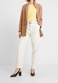 Žena v camel saku, žlutém topu, krémových kalhotách s páskem a černých teniskách na podpatku, stojící na bílém pozadí.