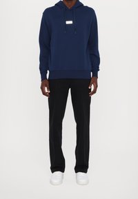 Navyblauer Hoodie mit Kordelzug und einem rechteckigen Logo-Patch auf der Brust, kombiniert mit schwarzer Hose und weißen Sneakers. Glatte Stoffstruktur.