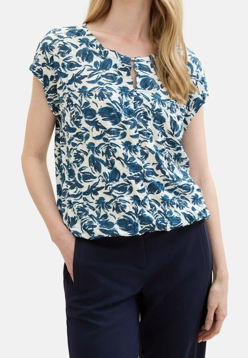 Femme portant un chemisier blanc à manches courtes avec un motif floral bleu et un pantalon bleu foncé, une main dans la poche, sur fond blanc.