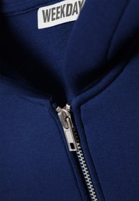 Μπλε ναυτικό φούτερ με φερμουάρ, διαθέτει μεταλλικό φερμουάρ και μαλακό, ελαστικό ύφασμα. Η ετικέτα  είναι ορατή μέσα στον γιακά και γράφει "WEEKDAY."