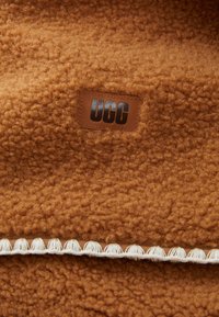 Fausse fourrure brune et texturée avec un patch logo UGG en cuir visible. Présente une bordure décorative blanche le long du bord.