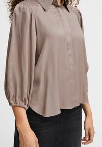 Helle braune Bluse mit Kragen, Ballonärmel und gerafften Bündchen. Der Stoff wirkt glatt mit vertikalen Texturdetaillierungen.