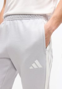 Άτομο που φοράει ανοιχτό γκρι παντελόνι φόρμας Adidas με ελαστική μέση και χέρι στην τσέπη, δείχνοντας το λογότυπο και τις τρεις ρίγες στο πλάι.