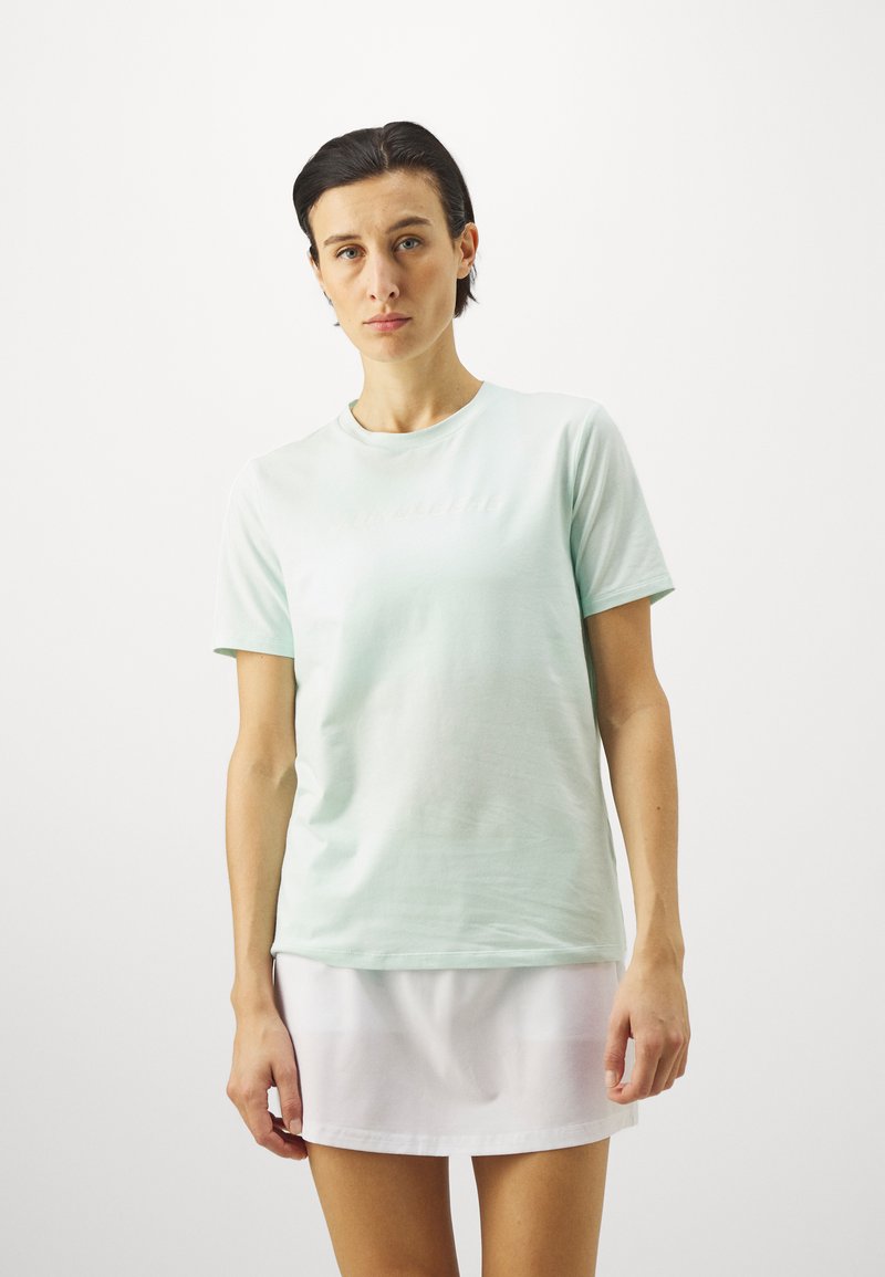 Camiseta de algodão verde menta clara com decote redondo e mangas curtas, usada com uma saia branca. Apresenta uma discreta marcação na parte da frente.