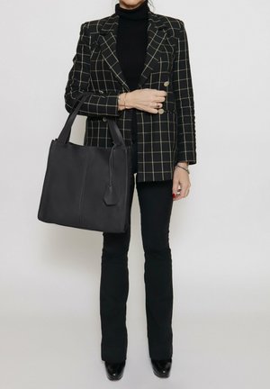 Zwarte leren tote bag met gestructureerd ontwerp, platte handvatten en een bijgevoegde label, gecombineerd met een geruite blazer en zwarte broek.