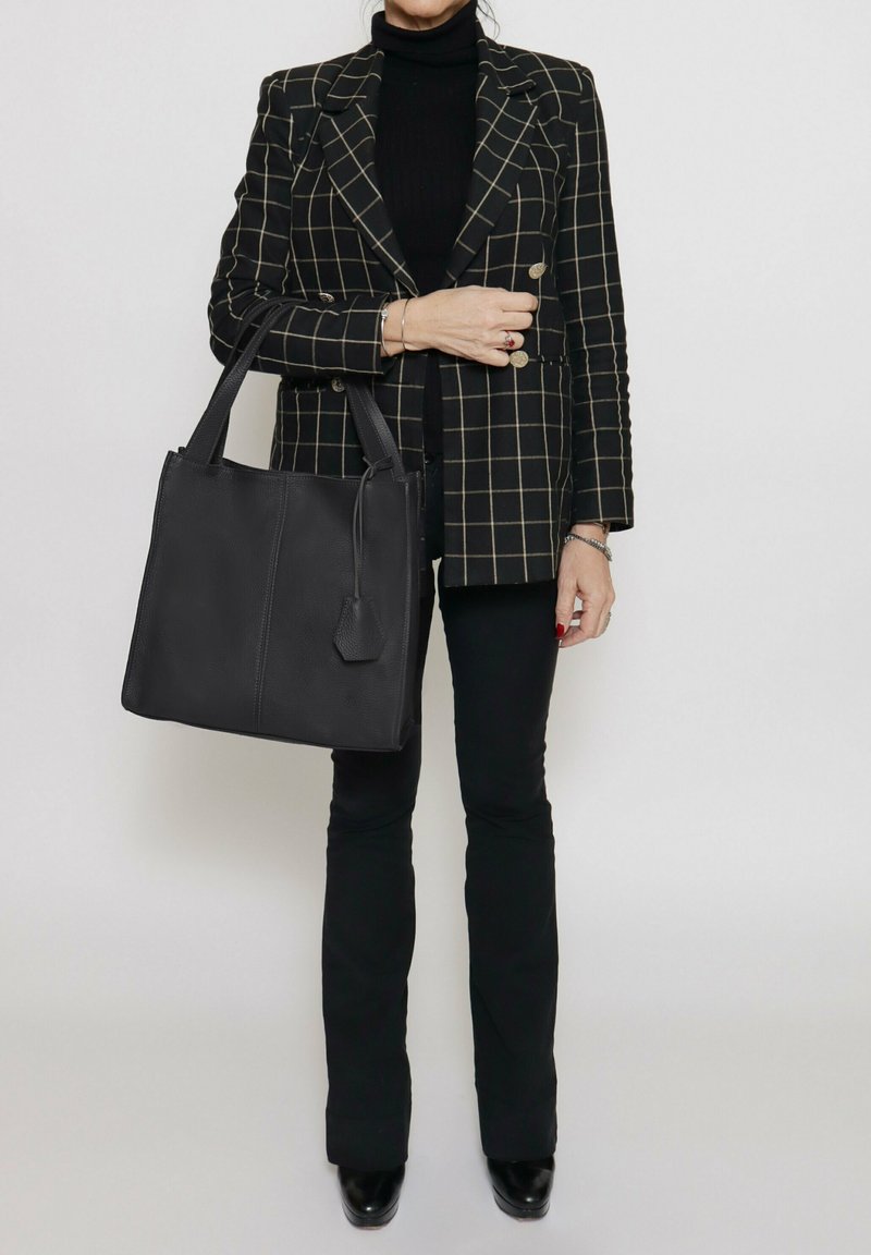 Borsa a tracolla nera in pelle con design strutturato, manici piatti e un'etichetta attaccata, abbinata a un blazer a scacchi e pantaloni neri.