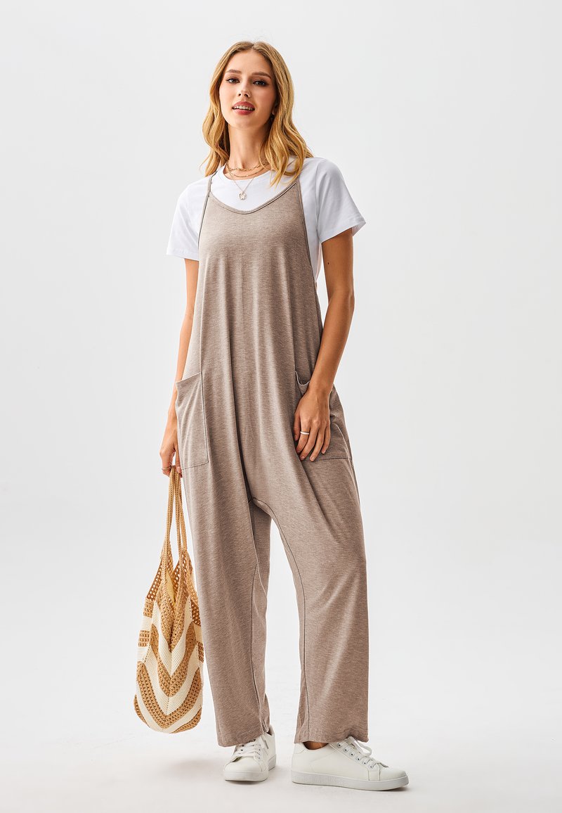 Beiger Jumpsuit mit weitem Bein, zwei Taschen vorne und Spaghettiträgern über einem weißen T-Shirt. Kombiniert mit weißen Turnschuhen und einer gemusterten Tragetasche.