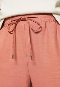 Teplákové kalhoty broskvové barvy vyrobené z texturované látky se elastickým pasem a šňůrkou na stažení s kovovými konci.