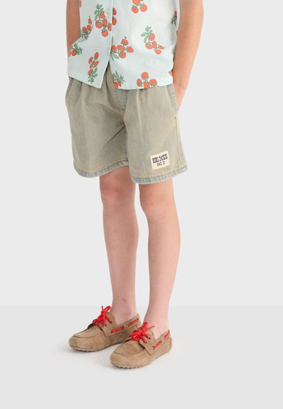 Child wearing beige shorts with a patch, a light shirt with orange fruit prints, and tan moccasins with red laces, standing with hands in pockets.