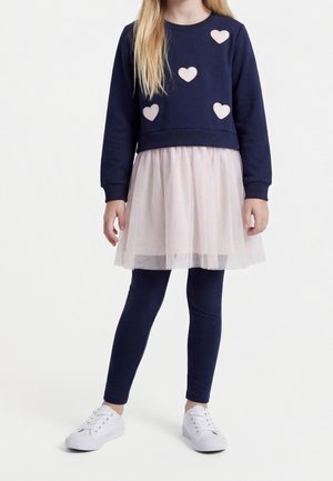 Fille portant un pull bleu marine avec des cœurs roses, une jupe en tulle rose clair par-dessus, des leggings bleu marine et des baskets blanches, debout sur fond blanc.