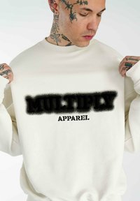 Cremefarbener Sweatshirt mit einem großen, schwarz gesprühten "MULTIPLY APPAREL"-Grafik in der Mitte. Baumwollmaterial mit gerippten Bündchen.