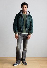 Grüne Satin-Bomberjacke mit grauem Futter, gerippten Bündchen und einem Logopatch. Kombiniert mit einem hellgrünen Strickpullover und grauer Jeans.