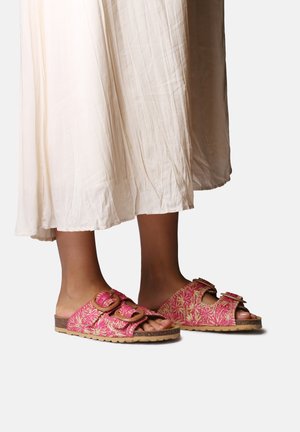 Różowe klapki na płaskim obcasie z dwoma regulowanymi paskami, teksturowaną powierzchnią i brązową podeszwą. Noszone z lekką białą spódnicą.