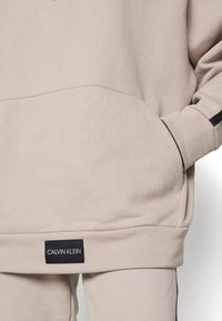 Sweatshirt beige avec une poche avant, texture douce, poignets côtelés et étiquette logo Calvin Klein noire sur l'ourlet.