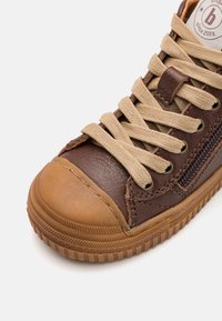 Hnědé kožené tenisky s tan-gumovým špičkovým krytem, béžovými tkaničkami, bočním zipem a texturovanou podrážkou na bílém pozadí.