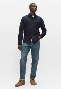 Marineblaue, strukturierte Hemdjacke mit Brusttasche, kombiniert mit hellblauen Jeans und braunen Wildlederstiefeln. Schlicht, entspannter Schnitt.