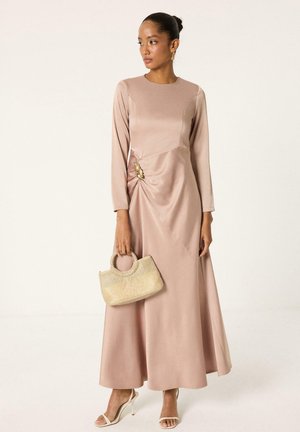 Femme portant une robe midi rose blush à manches longues avec un détail de broche dorée, tenant un sac à main tissé beige et portant des talons à brides.