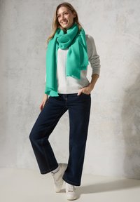 Turkisribbede tørklæde båret over en lys sweater, parret med mørke brede jeans og hvide sneakers, sat mod en tekstureret baggrund.