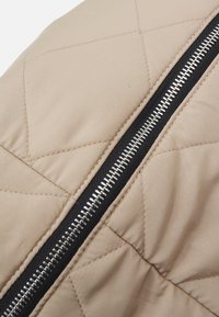 Gepolsterte beige Jacke aus Stoff mit einem schwarzen Reißverschluss. Die Textur ist glatt und weist rautenförmige Steppmuster auf. Silberne Reißverschluss-Beschläge sind sichtbar.