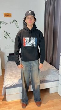 Młody mężczyzna ubrany w czarną bluzę z kapturem z kolorowym nadrukiem w czerwonym, białym i niebieskim kolorze, szare dżinsy, brązowe buty oraz ciemną czapkę z daszkiem, stoi w sypialni.